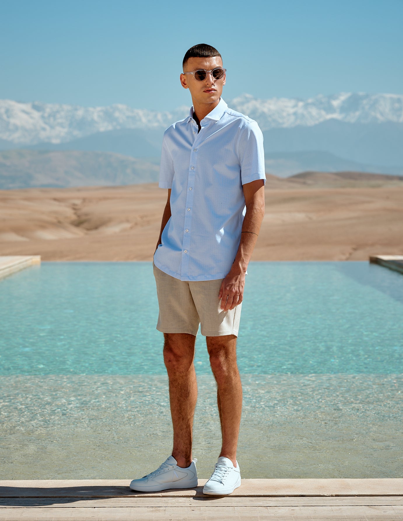 Male model wearing beige shorts, light blue short-sleeved shirt and sunglasses standing in a nature summer setting