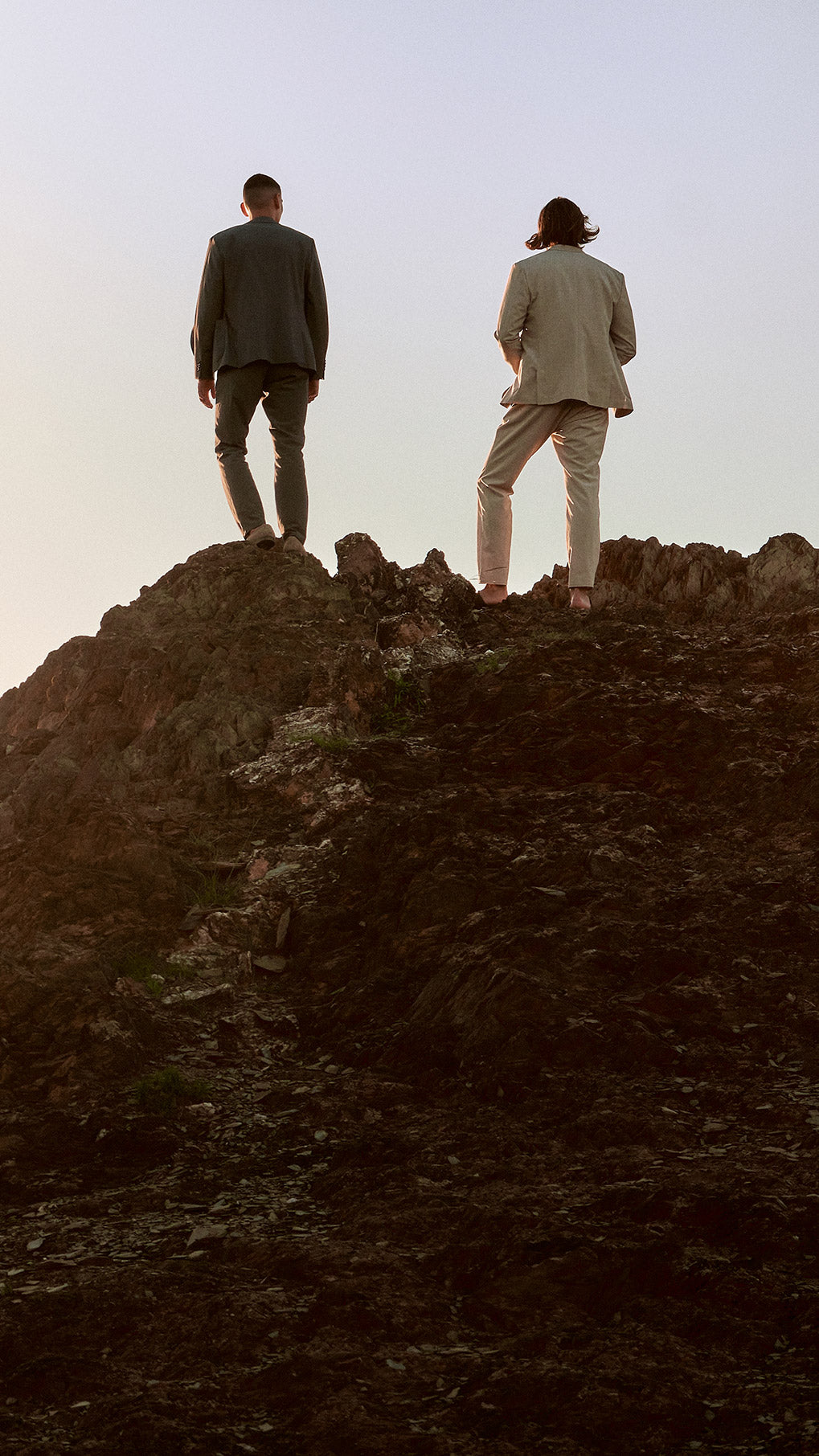 Two men standing on the top of the mountain looking into the distance and wearing a grey and beige suit.