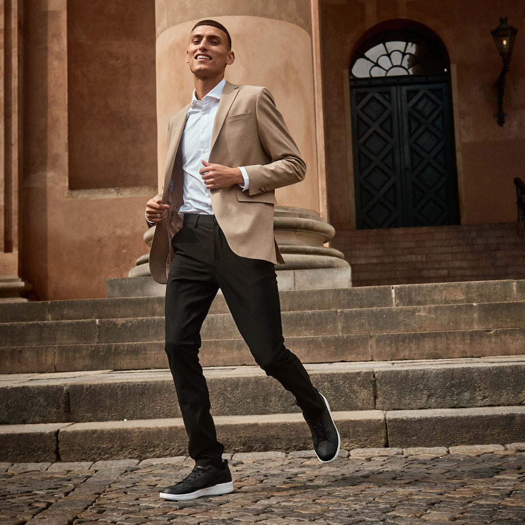 Man running in front of urban building wearing black pants, white shirt and beige blazer