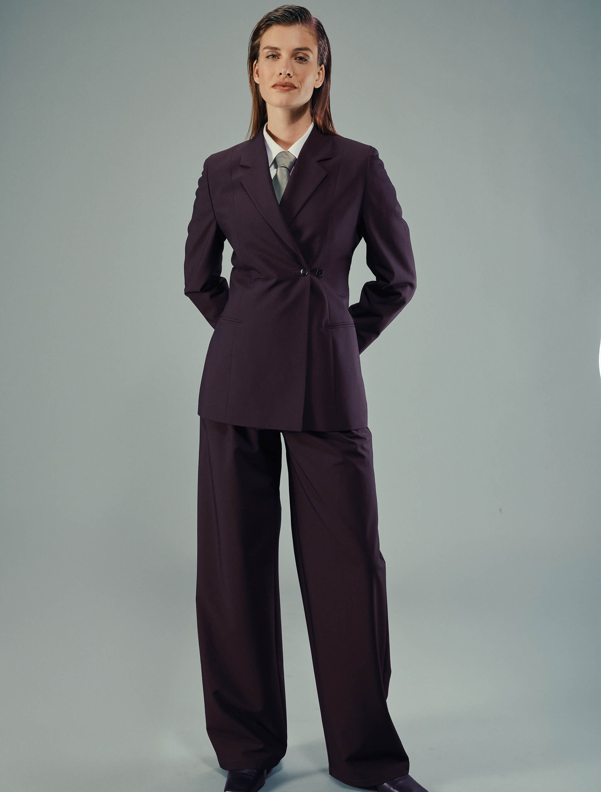 A person wearing a Women’s Avira Blazer Burgundy Berry with matching wide-leg trousers, styled with a white collared shirt and a grey tie in a refined Scandinavian design.