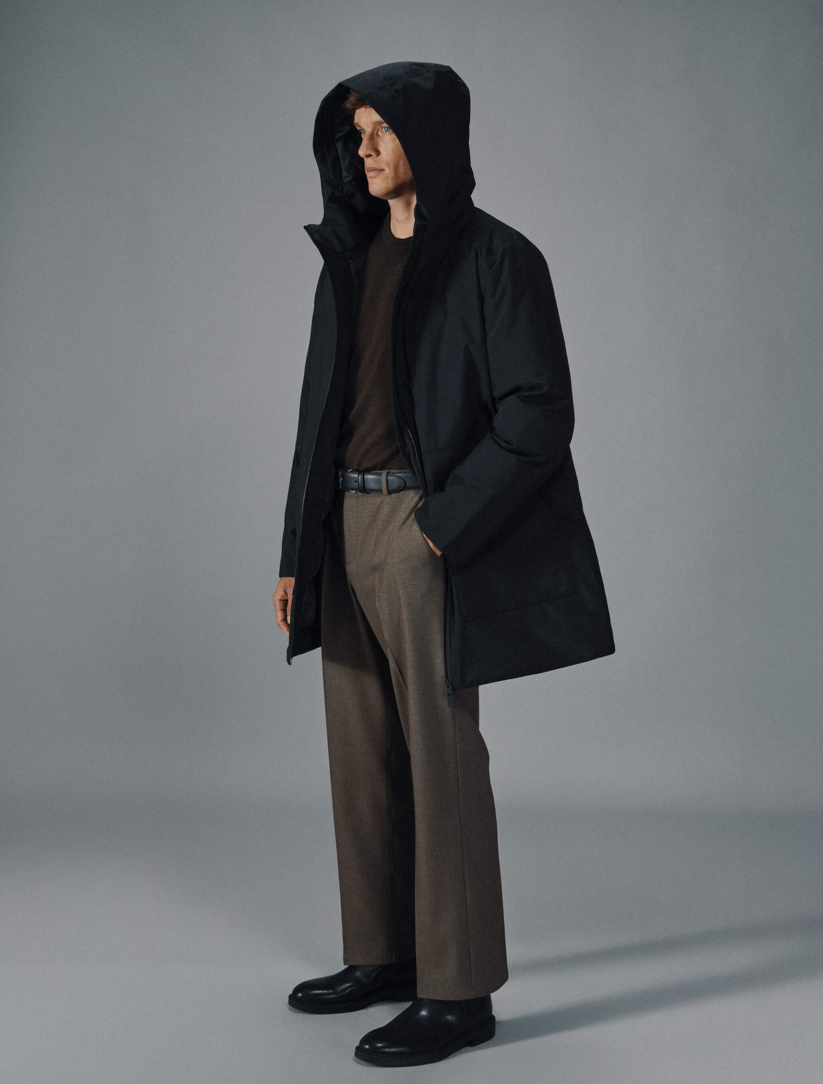 A man wears a black Utility Jacket Black over a dark brown sweater, brown trousers, and black shoes.