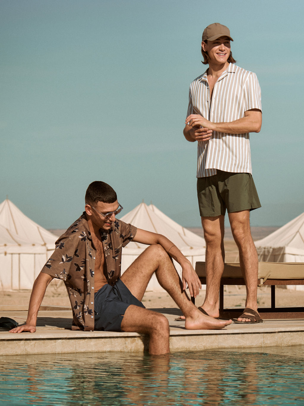 Two male models wearing different swim shorts and short-sleeved shirts by a pool