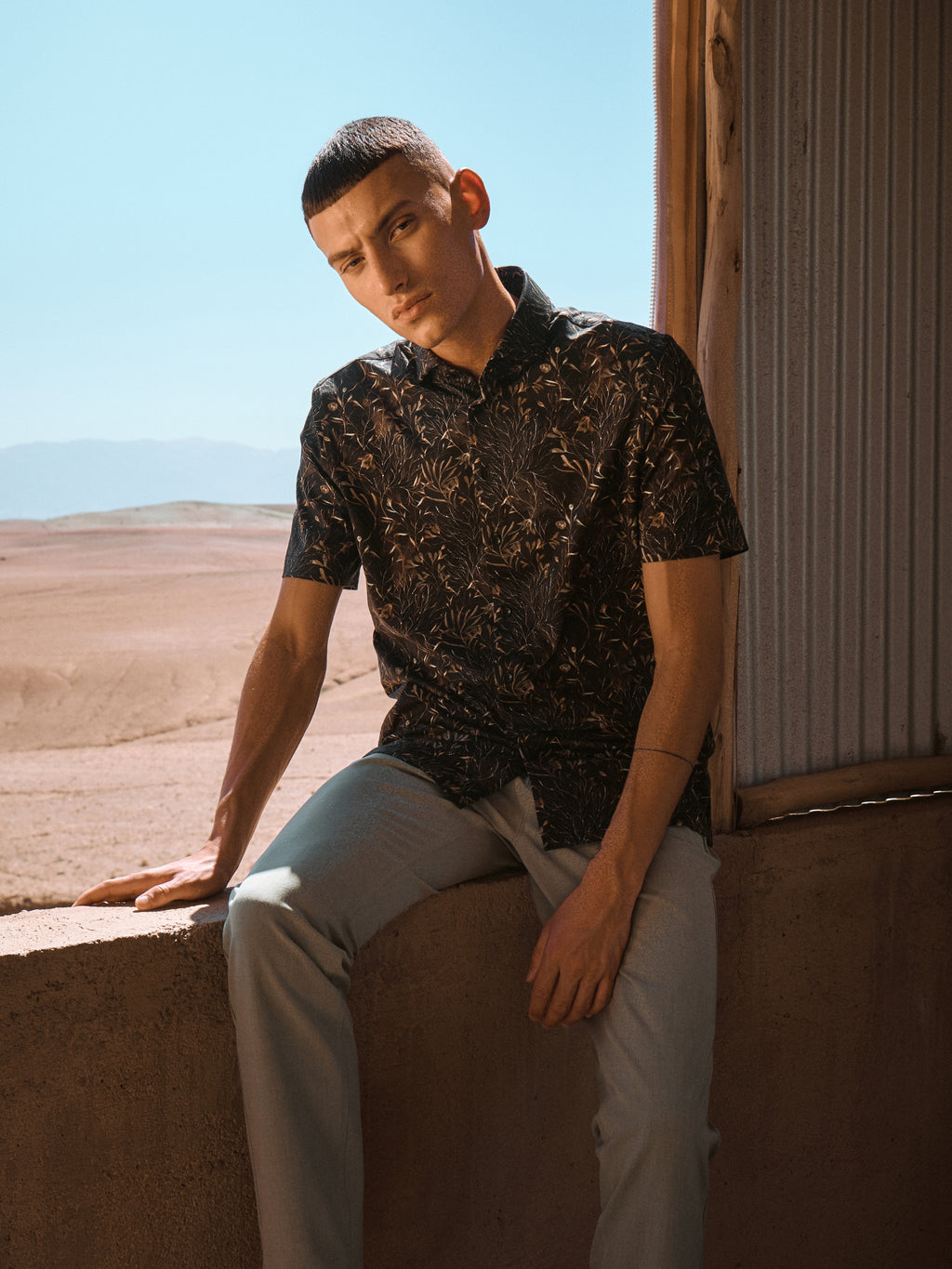 Male model wearing dark patterned short-sleeved shirt and beige pants in a summer nature setting
