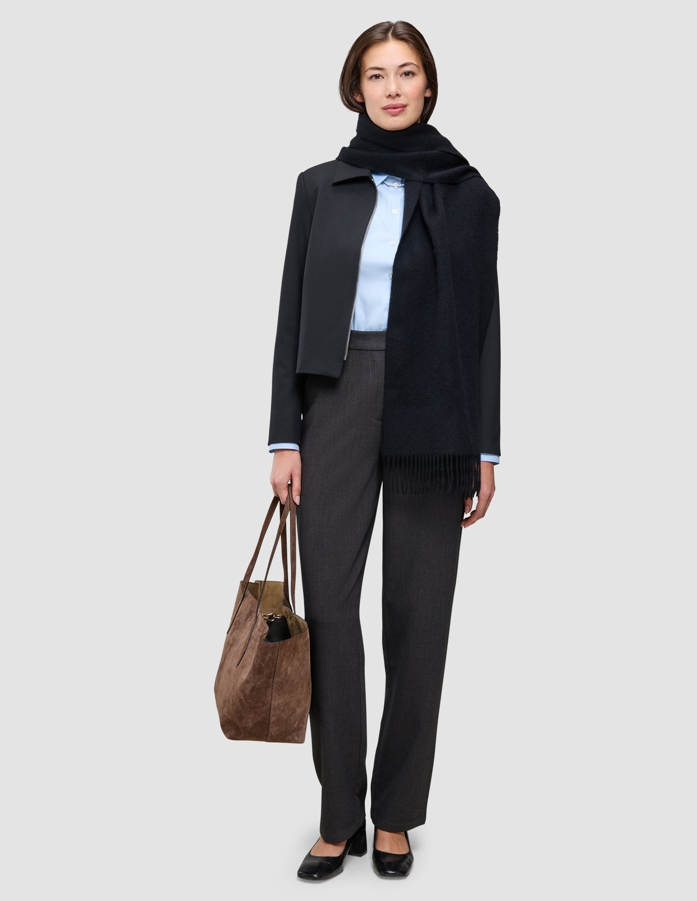 A woman wears an An easy office upgrade over a light blue Oxford shirt, dark trousers, and a black scarf, carrying a brown tote bag.