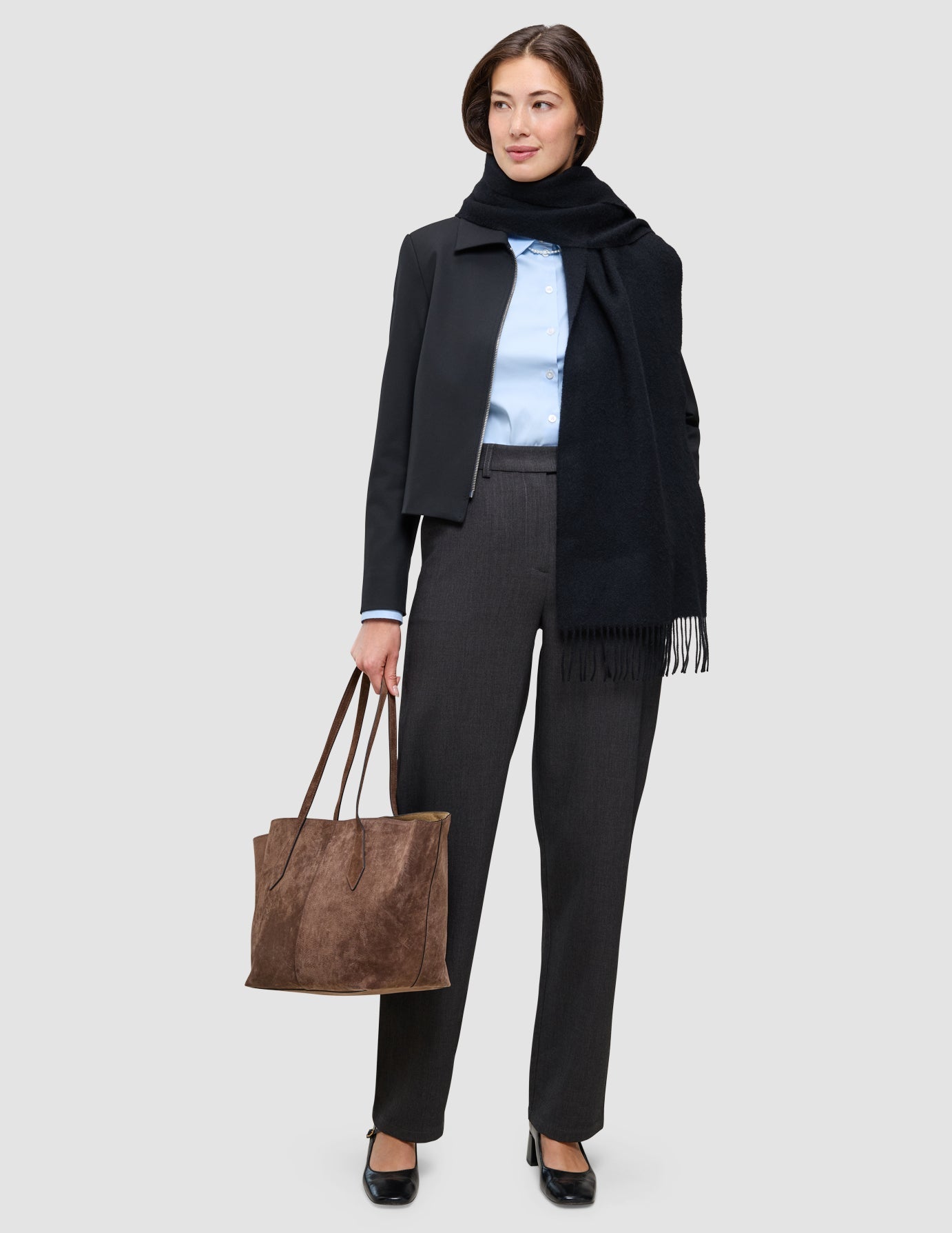 An easy office upgrade: a person wearing a black Essential Zip Jacket over a light blue button-down shirt, charcoal Go2 Pants, and a black scarf, holding a large brown tote bag.