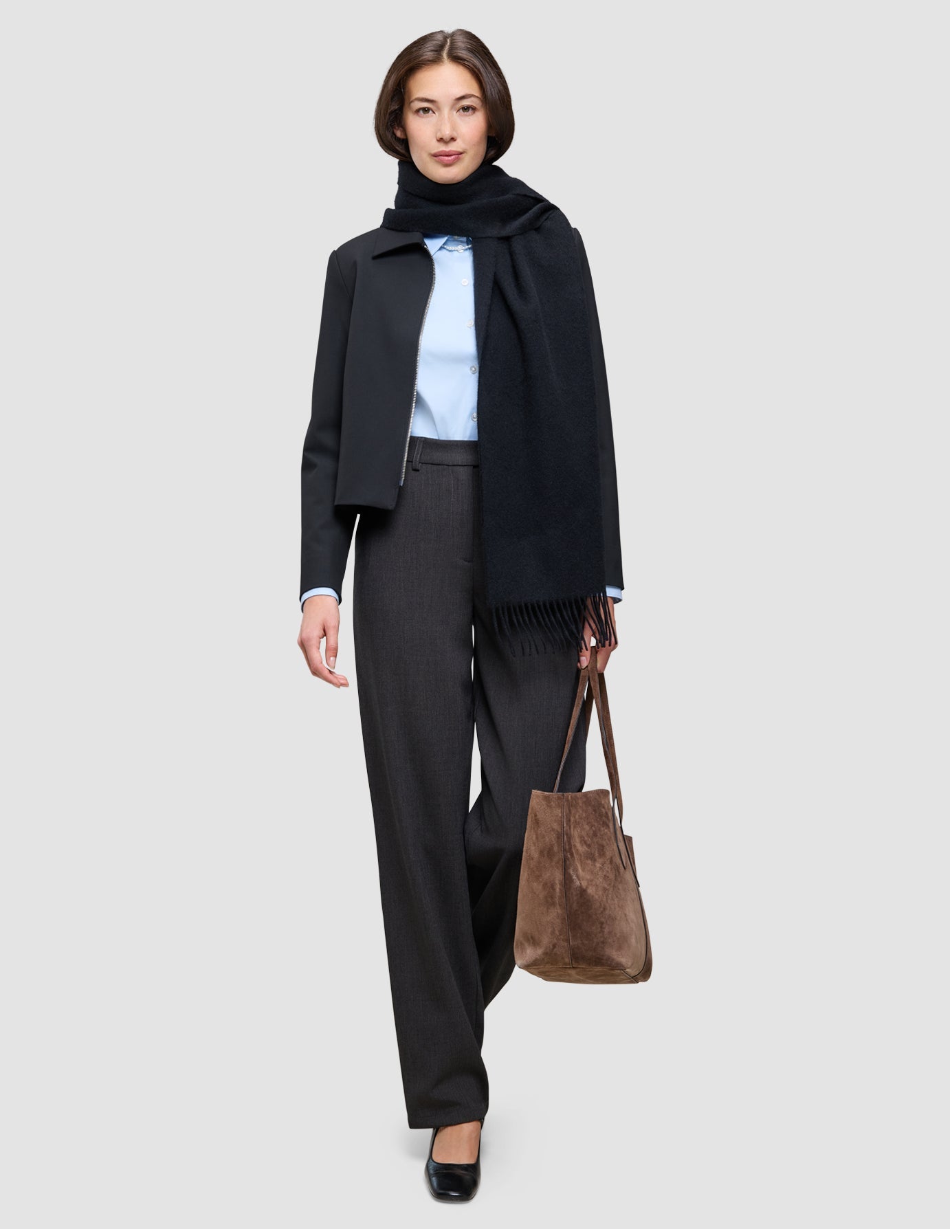 An easy office upgrade: a person wearing a light blue Oxford shirt, dark trousers, a short dark jacket, and a long dark scarf, holding a brown suede tote bag.
