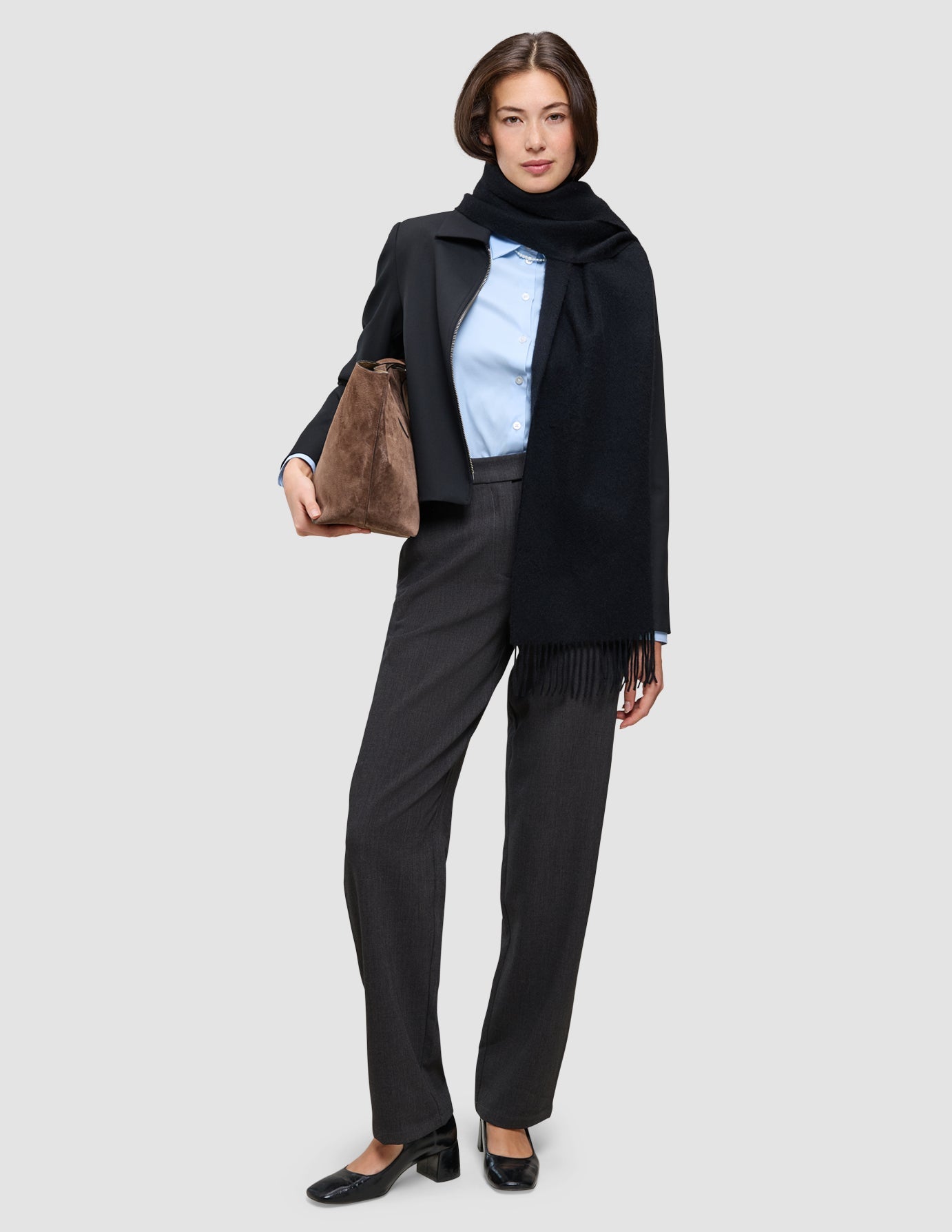 A woman wearing an An easy office upgrade over a light blue Oxford shirt, charcoal trousers, and a black scarf, holding a brown handbag.