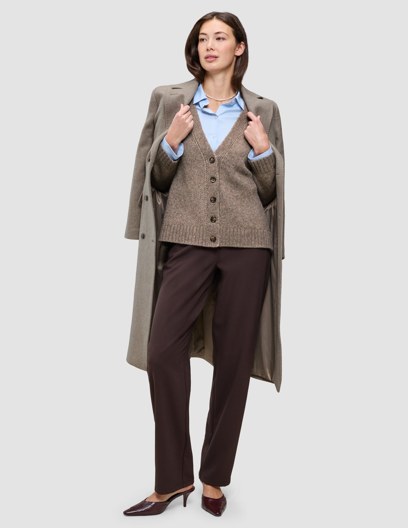 A woman wearing Cold-weather workwear consisting of a tan wool coat draped over her shoulders, a brown button-front cardigan, a light blue oxford shirt, and brown trousers.