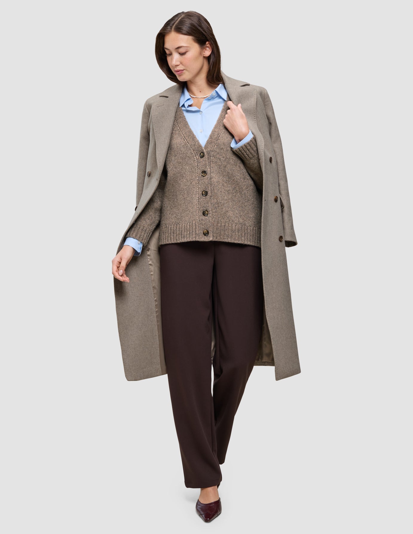 A person wearing Cold-weather workwear, consisting of a light blue Oxford shirt, a brown v-neck cardigan, dark Go2 pants, and a long coat draped over their shoulders.