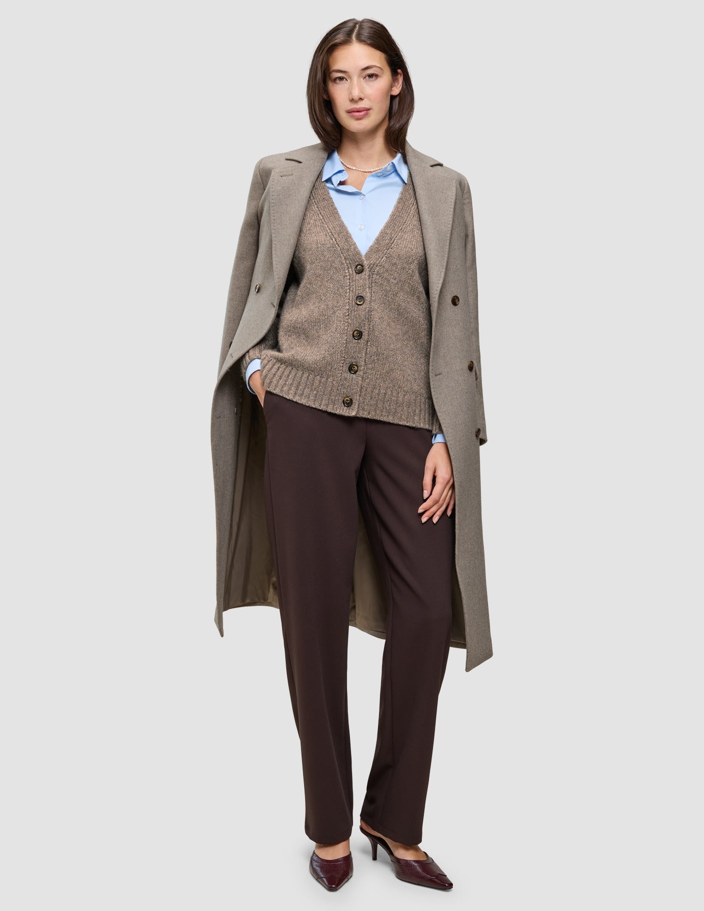 A person wearing Cold-weather workwear, consisting of a light brown wool coat over a brown knit cardigan, a blue Oxford shirt, and dark Go2 pants.