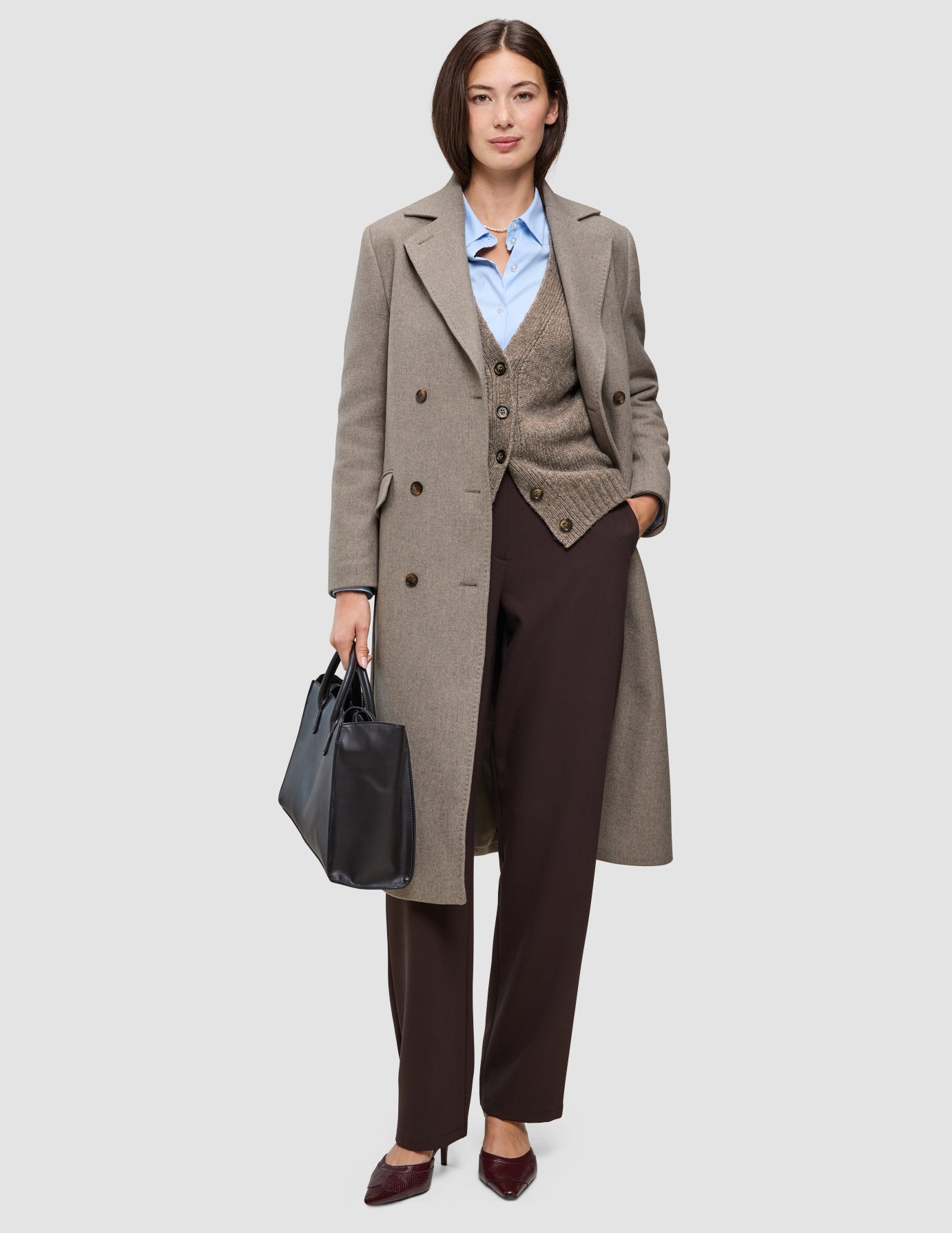 A woman wearing Cold-weather workwear, specifically a tan wool coat over a brown cardigan, blue Oxford shirt, and Go2 Pants, carrying a black tote bag.