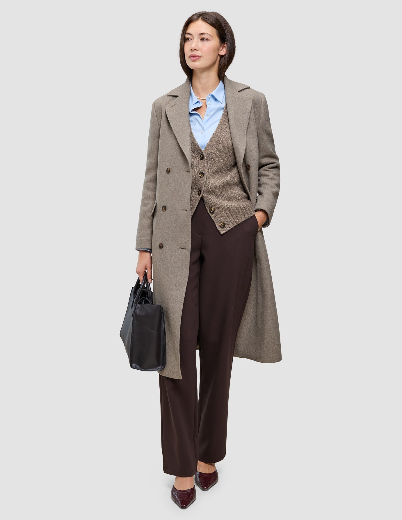 A woman wearing Cold-weather workwear, specifically a tan wool coat over a brown cardigan, blue Oxford shirt, and dark Go2 pants, holding a black briefcase.