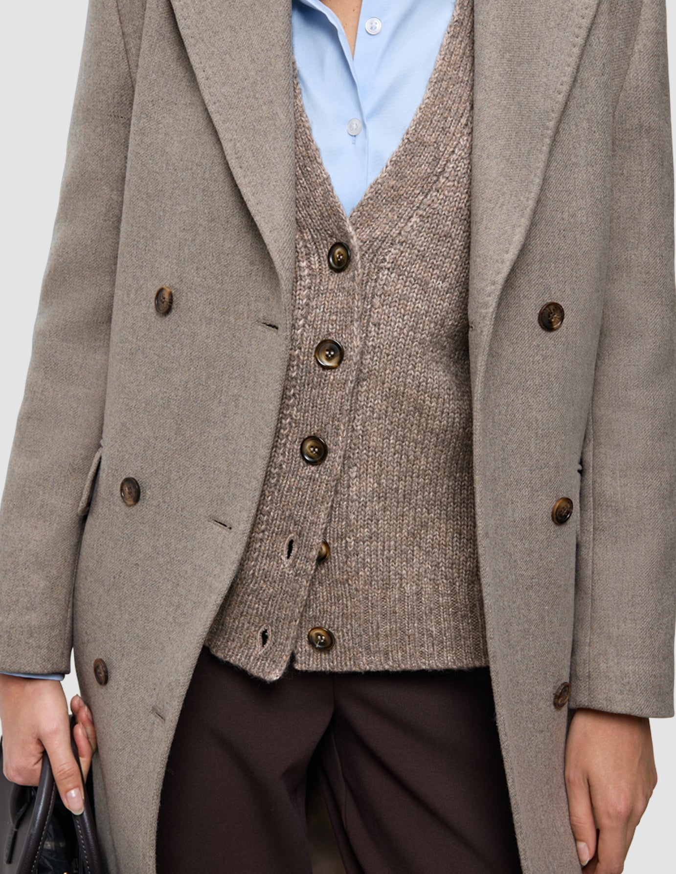 A person wears Cold-weather workwear consisting of a light blue Oxford shirt, a brown knitted cardigan, and a beige wool coat over dark brown Go2 pants.