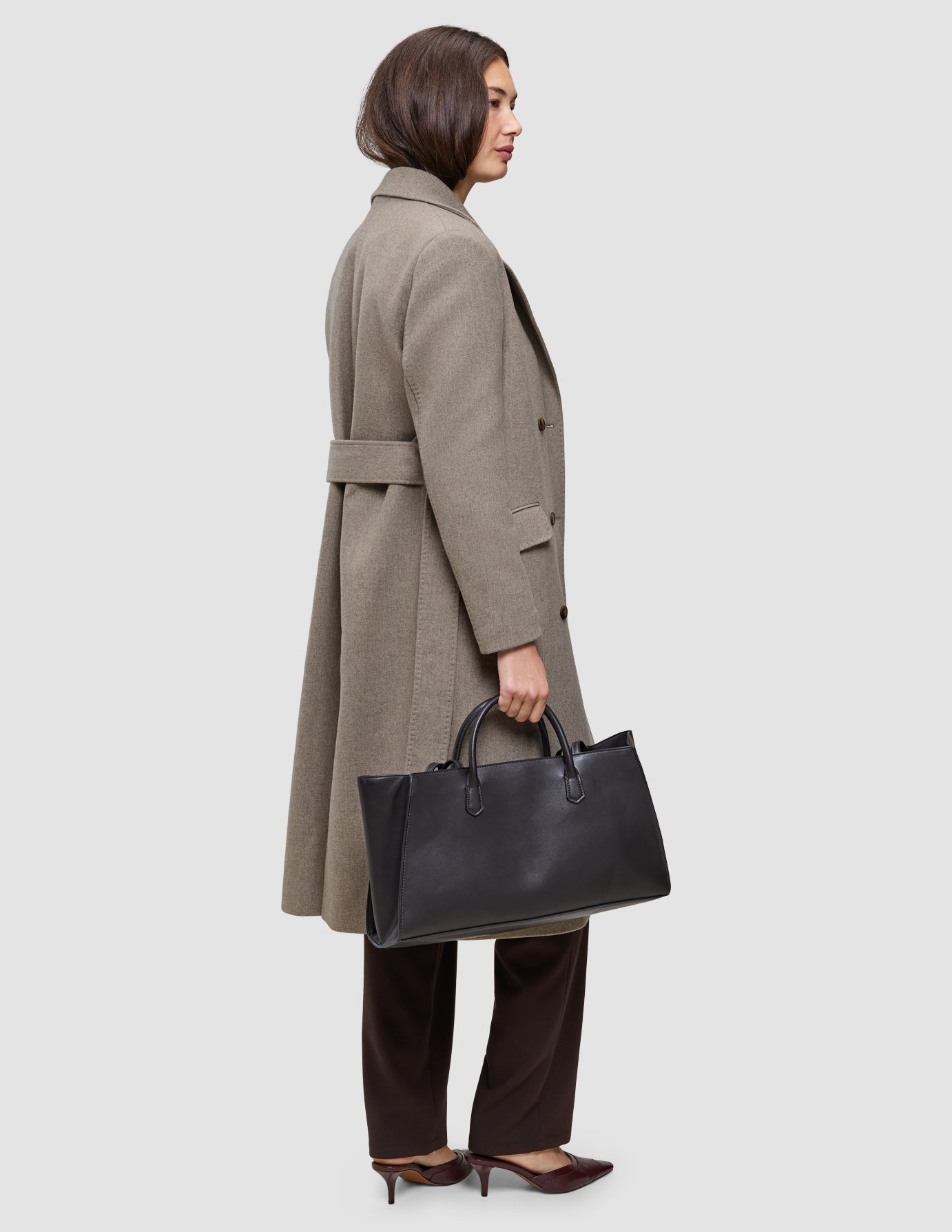 A person wearing Cold-weather workwear, specifically a long, belted taupe wool coat and dark Go2 pants, carries a black leather handbag.