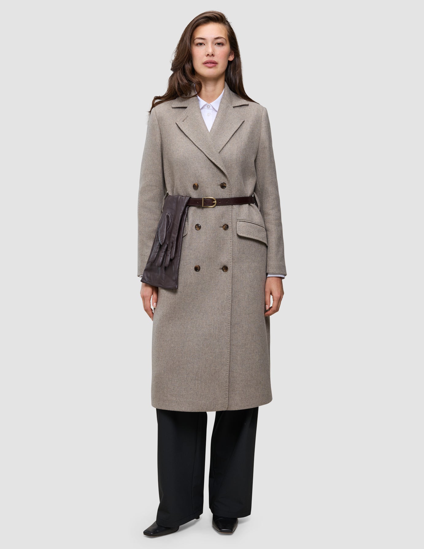 For Winter, dressed up, a person wears a beige double-breasted wool coat over a white Oxford shirt and black trousers, cinched with a dark leather belt holding a pair of brown gloves.