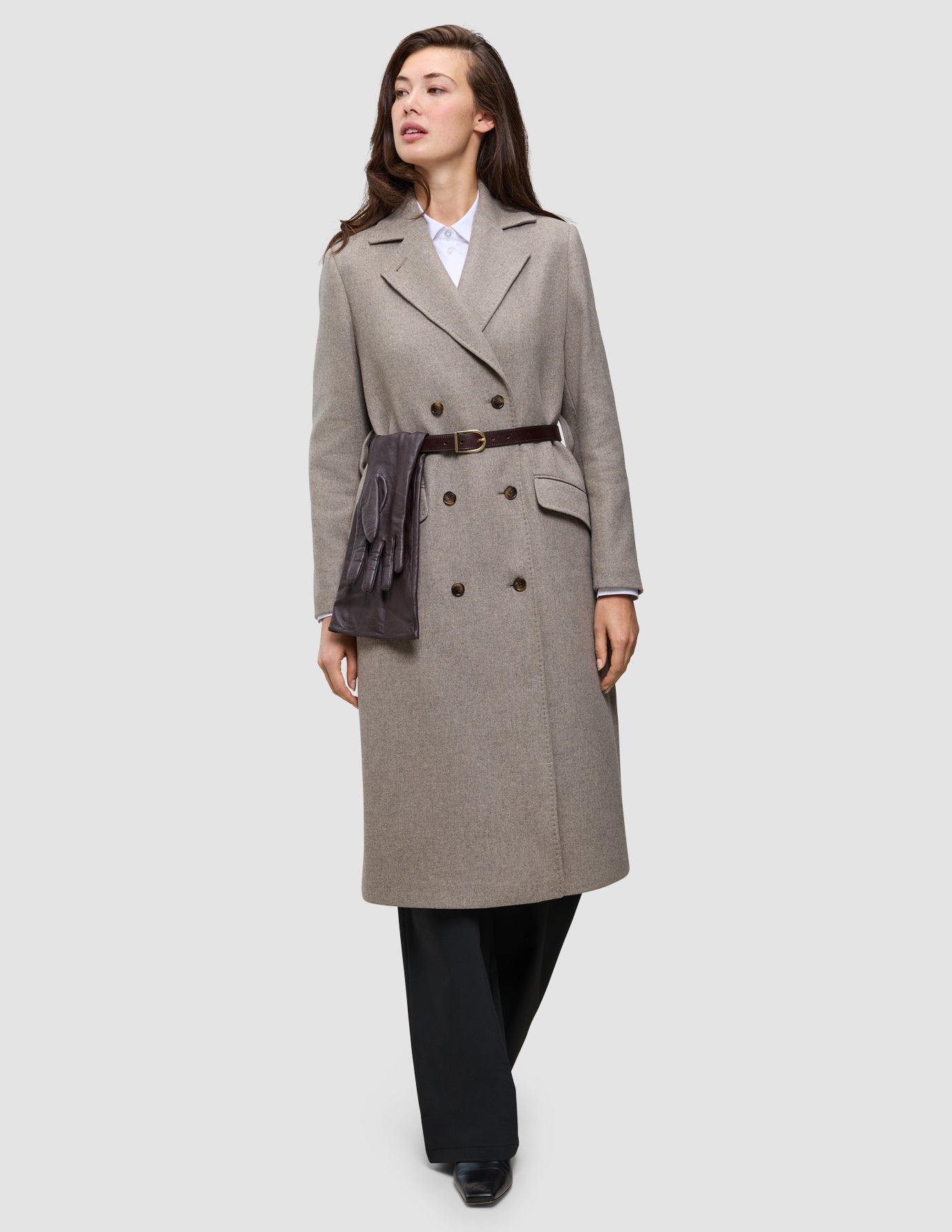 Winter, dressed up: a person wearing an Oxford shirt under a long, double-breasted taupe wool coat, belted at the waist with brown leather gloves tucked into the belt.