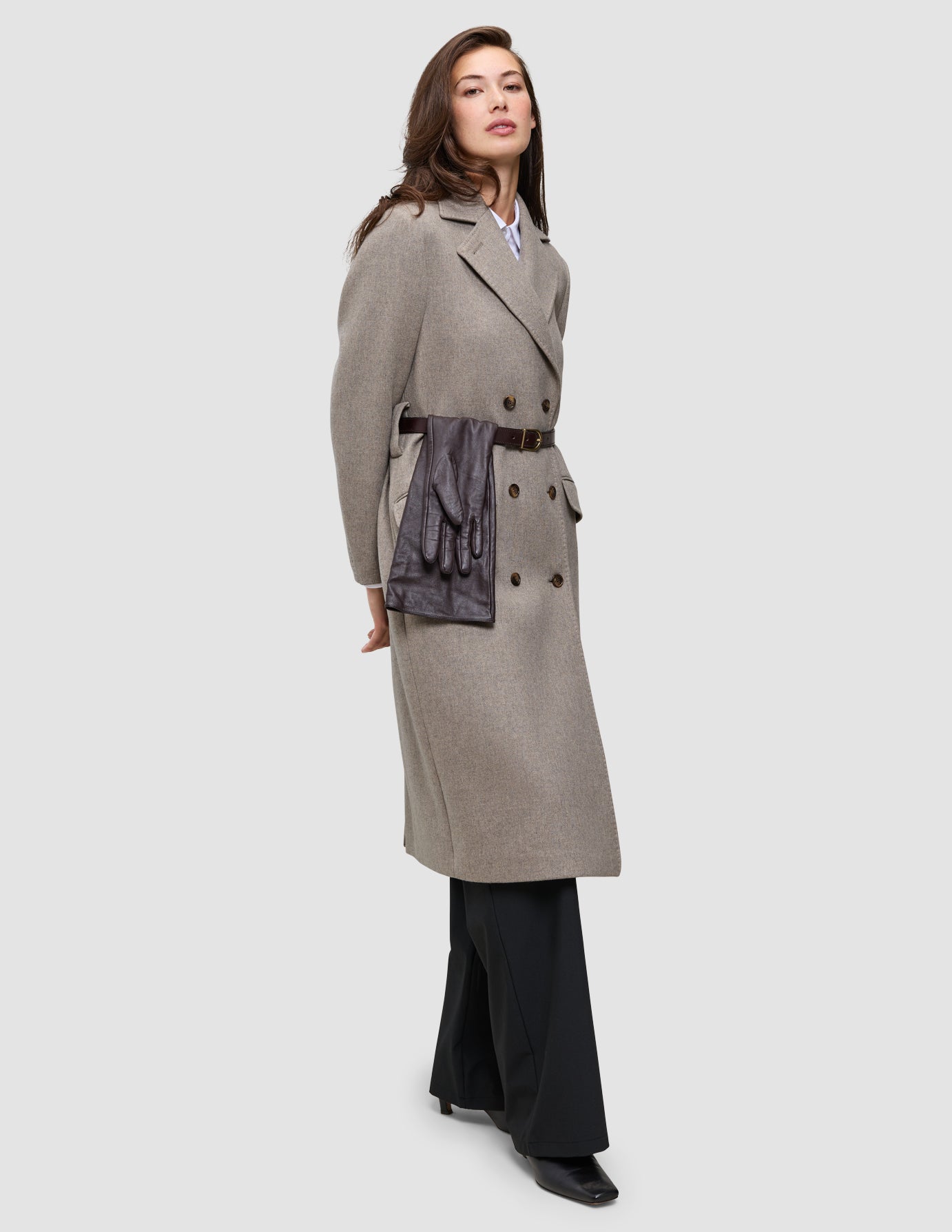 A person wears the Winter, dressed up beige double-breasted wool coat over an Oxford shirt and black trousers, with brown leather gloves tucked into a thin brown belt at the waist.