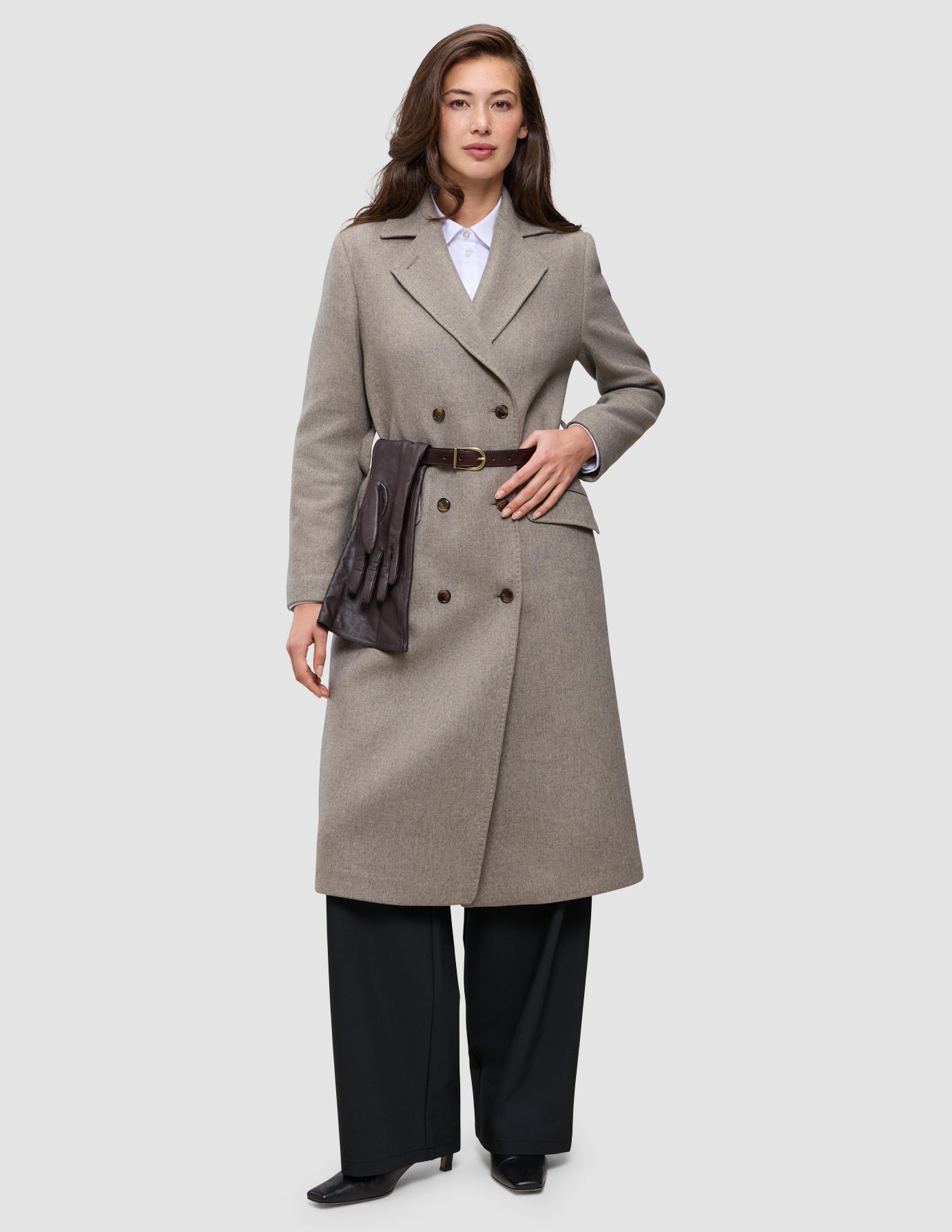 A woman in Winter, dressed up wears a beige double-breasted wool coat over a white Oxford shirt and black trousers, cinched with a dark leather belt holding a pair of brown gloves.
