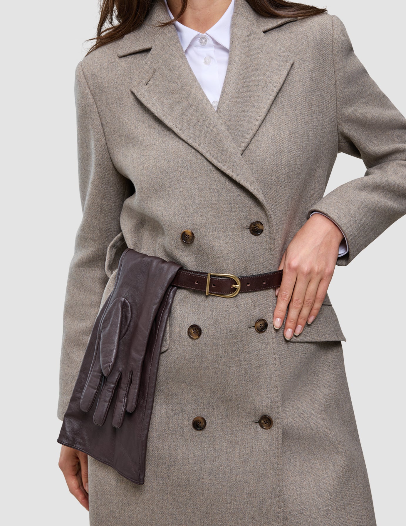 A person wearing the Winter, dressed up double-breasted wool coat over a white Oxford shirt, accessorized with a dark brown leather belt and matching gloves tucked into the waist.