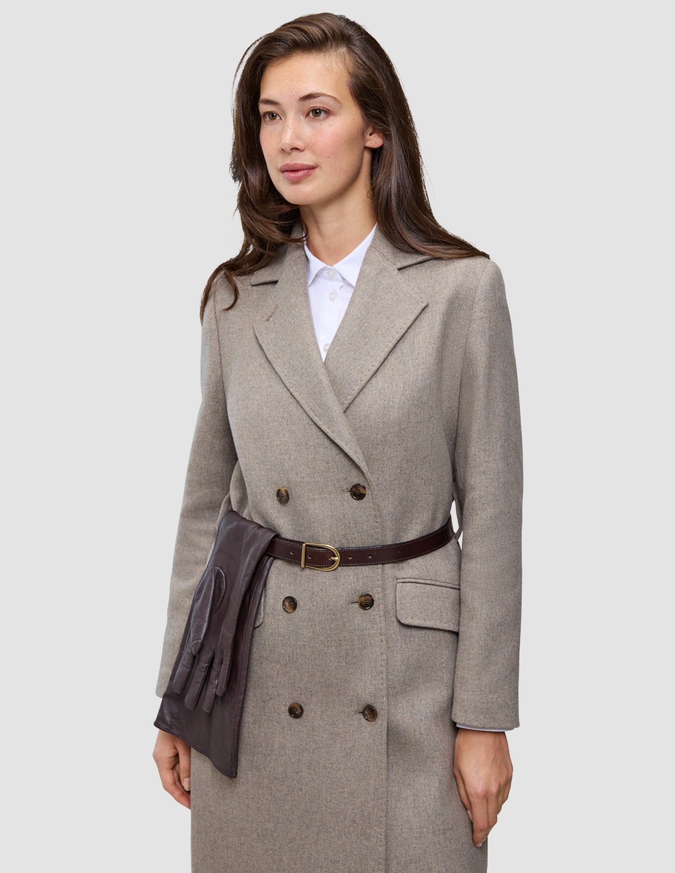 A woman wearing a Winter, dressed up double-breasted beige wool coat over a white oxford shirt, with a dark brown leather belt holding a pair of matching leather gloves at her waist.