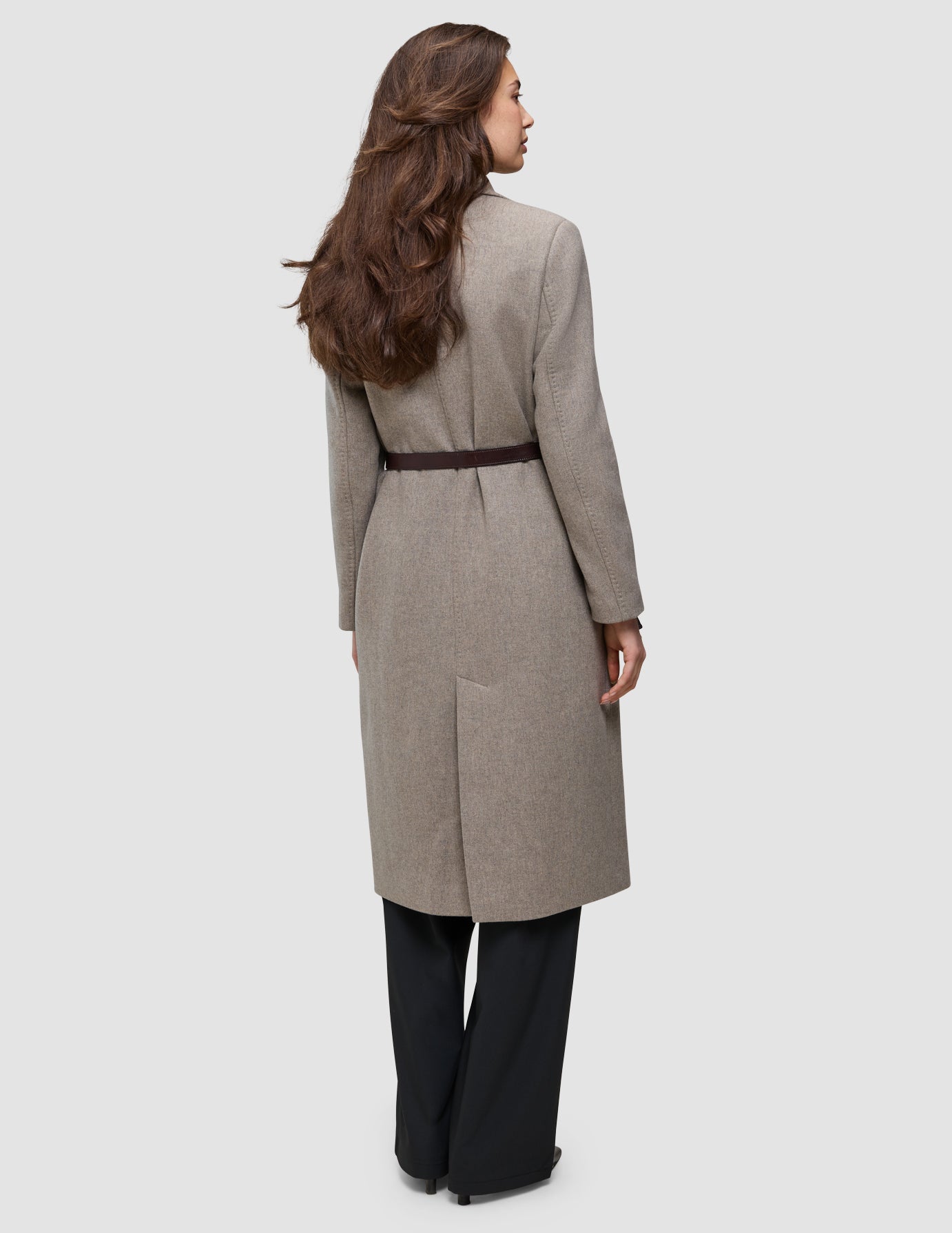 A woman from behind wears the Winter, dressed up tan, knee-length wool coat over an Oxford shirt, styled with a dark brown belt and black trousers.