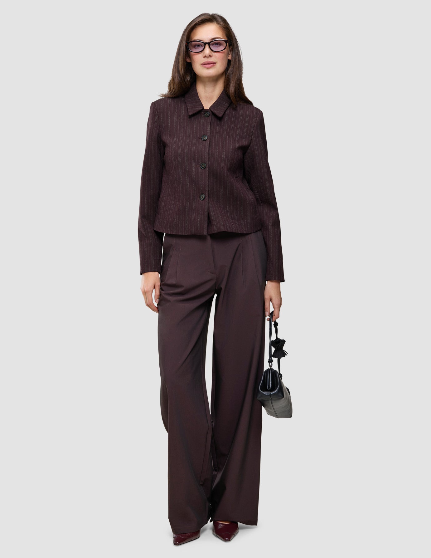 A woman wearing a Burgundy from top to toe dark burgundy jacket and wide-leg trousers, holding a small black handbag.