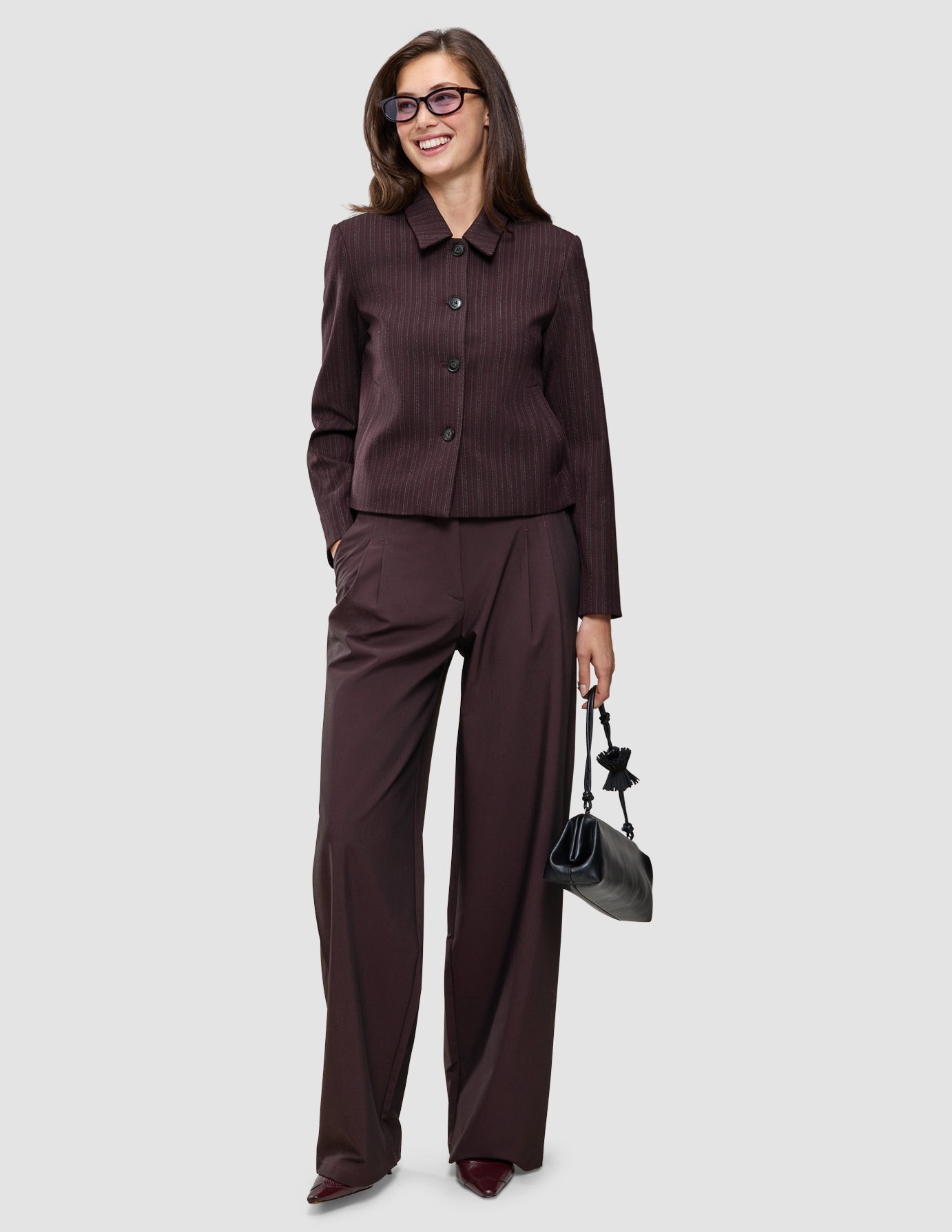 A smiling woman wears a Burgundy from top to toe outfit consisting of a dark burgundy pinstripe jacket and wide-leg trousers, holding a small black handbag.