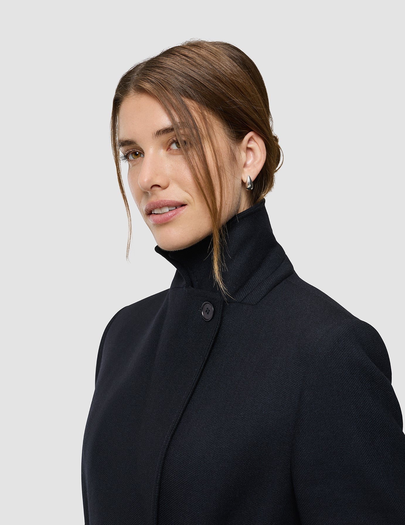 A woman wearing a black high-neck coat looks toward the camera.