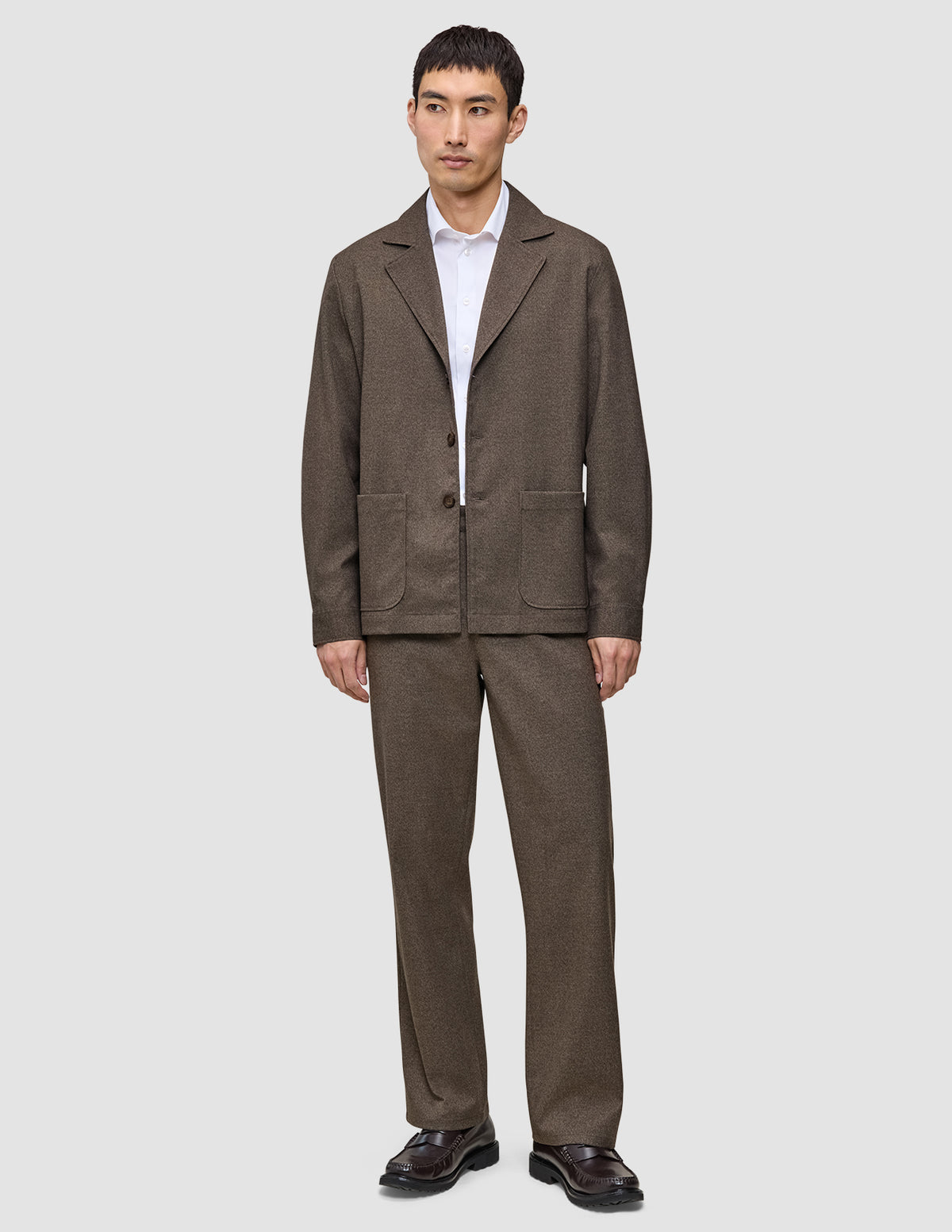 A person wearing an Atelier Overshirt Dark Beige with matching trousers over a white collared shirt, styled with dark loafers for a clean Scandinavian design aesthetic.