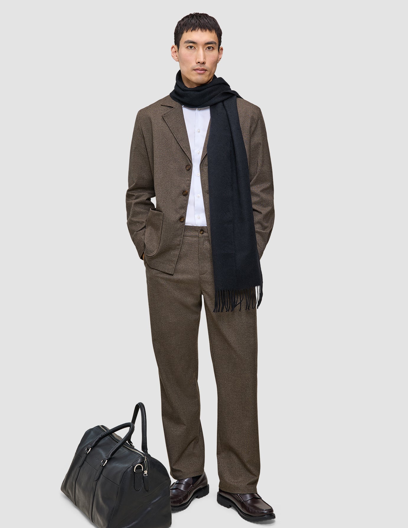A man wearing a dark beige Atelier Overshirt heavyweight twill blazer over a white shirt with a dark scarf stands next to a black leather duffel bag.