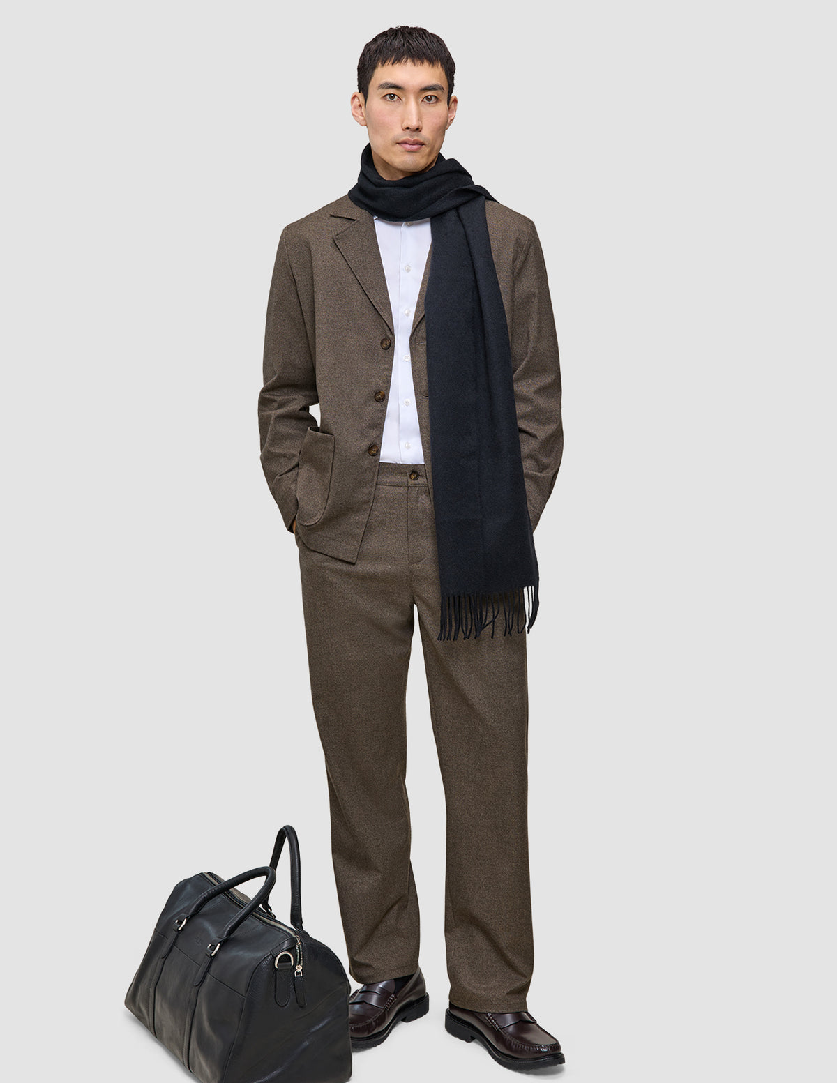 A man wearing a dark beige Atelier Overshirt heavyweight twill blazer over a white shirt with a dark scarf stands next to a black leather duffel bag.