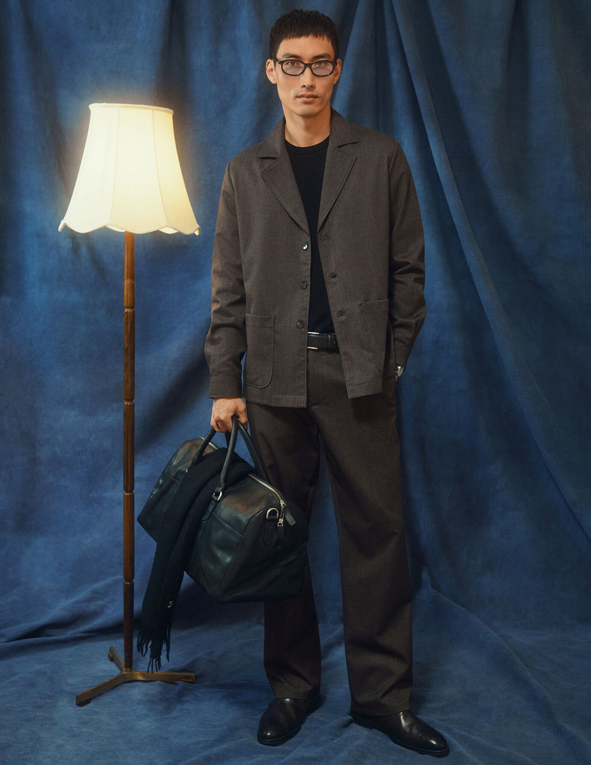 A man in a dark beige Atelier Overshirt, matching trousers, and a black shirt holds a dark leather duffel bag.