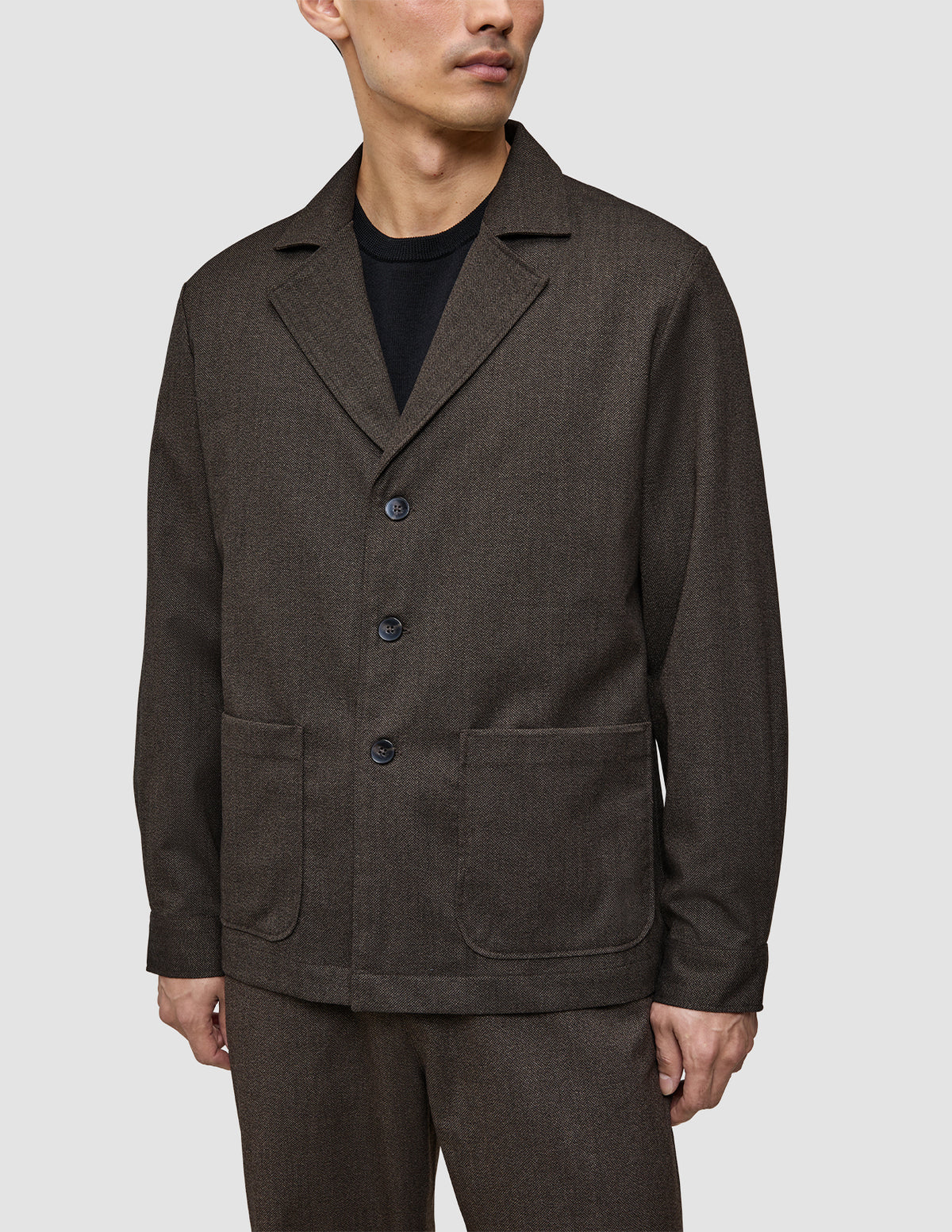 A person wearing an Atelier Overshirt Dark Brown, a Scandinavian-designed blazer with notch lapels, three buttons, and two large patch pockets.