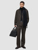 A man wearing an Atelier Pants Relaxed Dark Brown suit, a black sweater, and a scarf, while holding a black leather duffel bag.
