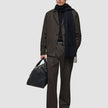 A man wearing an Atelier Pants Relaxed Dark Brown suit, a black sweater, and a scarf, while holding a black leather duffel bag.