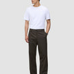 A person wearing a white t-shirt and Atelier Pants Relaxed Dark Brown in heavyweight broken twill, paired with dark leather shoes.