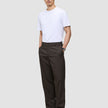 A man wearing a white t-shirt and Atelier Pants Relaxed Dark Brown in heavy-weight broken twill, standing with his hands in his pockets.