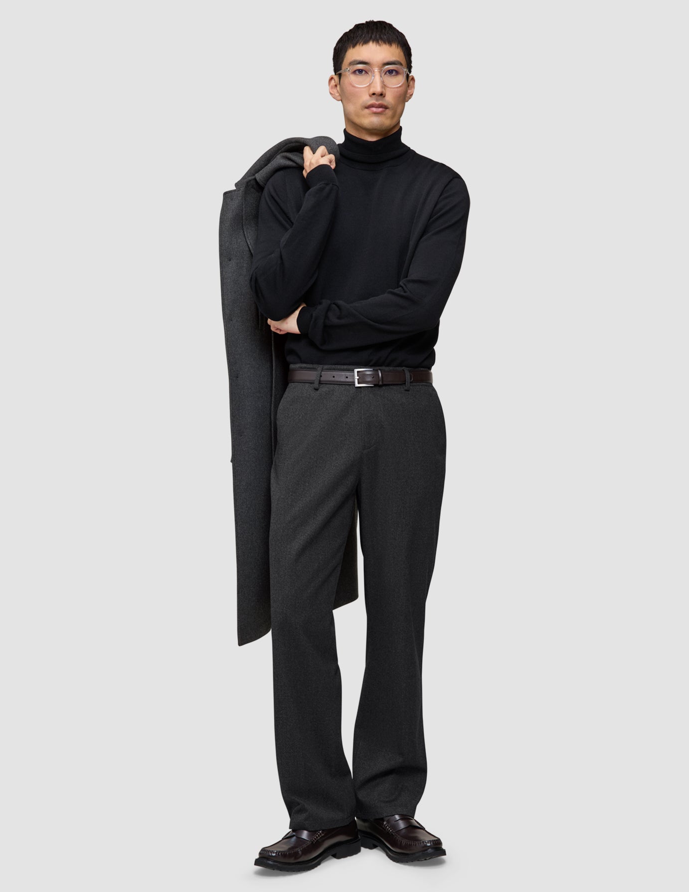 A man wearing a black turtleneck and Atelier Pants Relaxed Dark Grey in heavyweight broken twill, holding a coat over his shoulder.