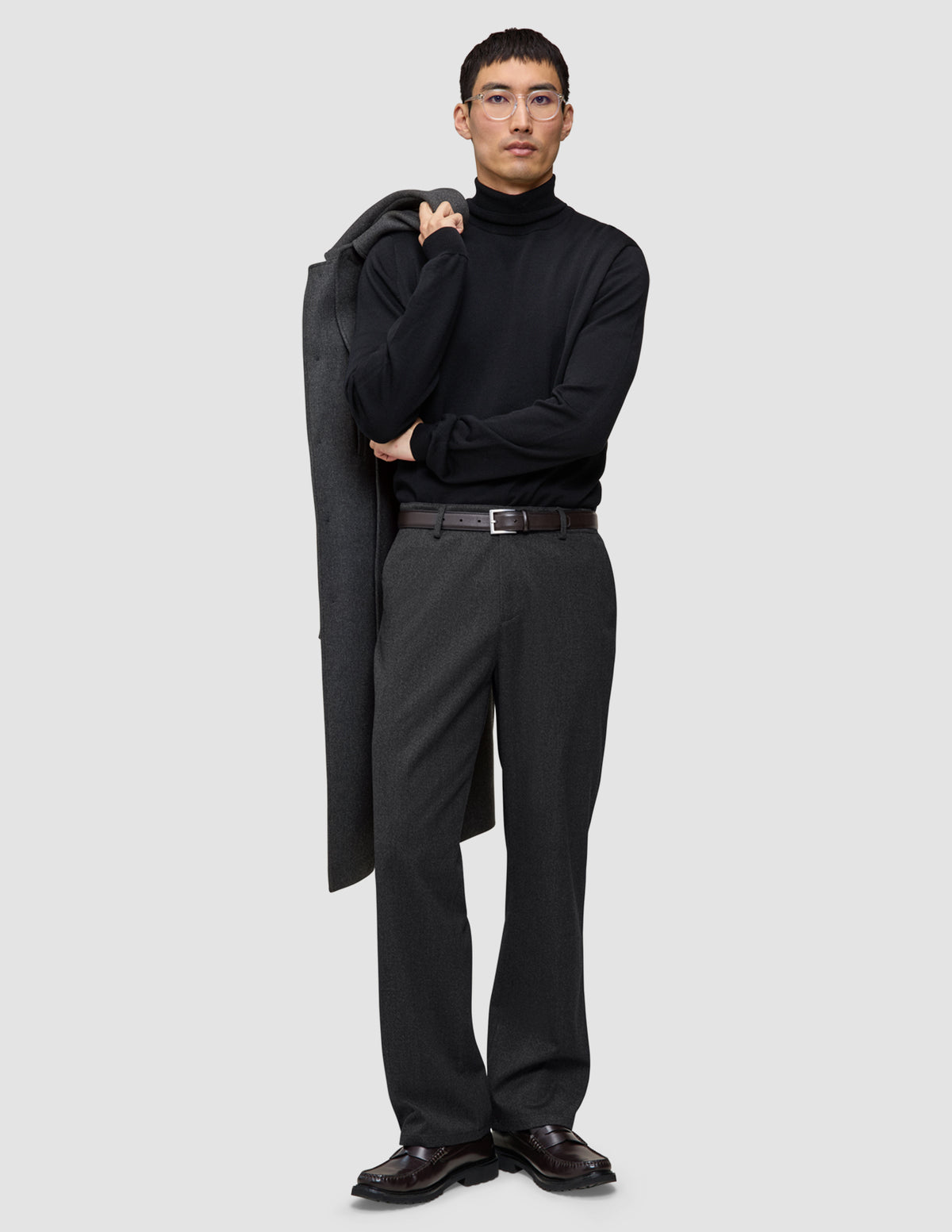 A man wearing a black turtleneck and Atelier Pants Relaxed Dark Grey in heavyweight broken twill, holding a coat over his shoulder.