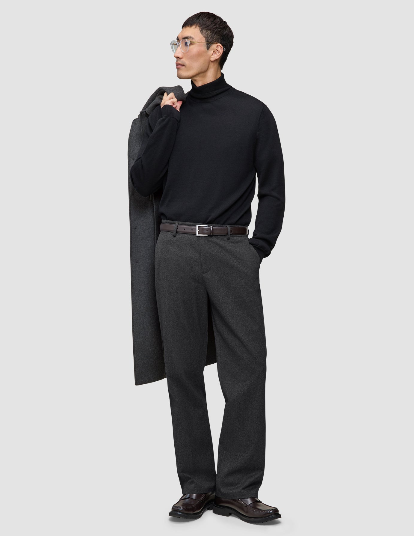 A man wearing a black turtleneck and Atelier Pants Relaxed Dark Grey in heavyweight broken twill, holding a coat over his shoulder.