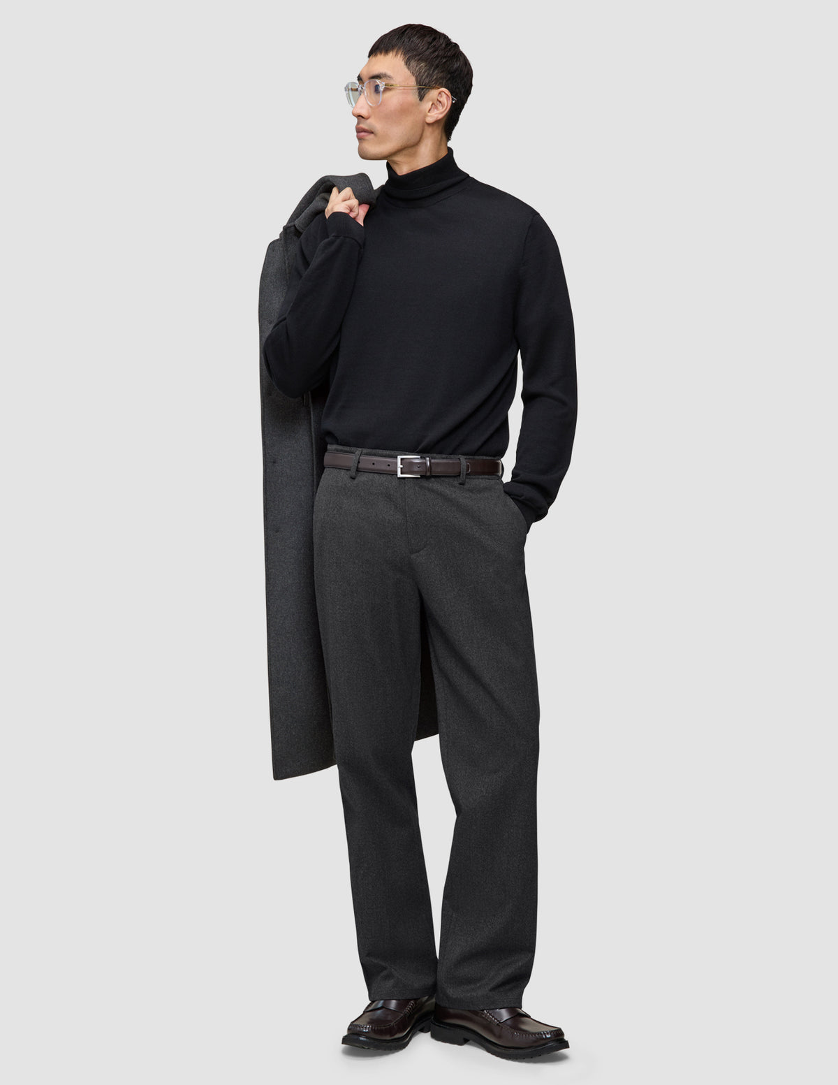 A man wearing a black turtleneck and Atelier Pants Relaxed Dark Grey in heavyweight broken twill, holding a coat over his shoulder.
