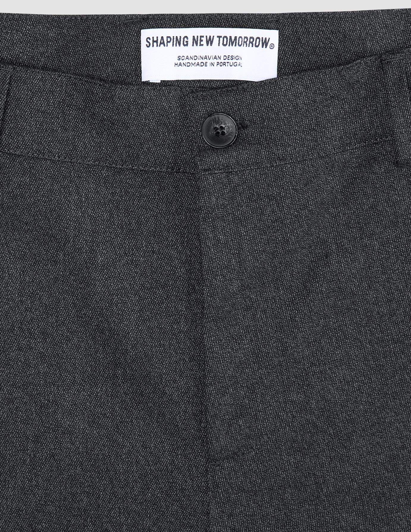 Close-up of the waistband and button of Atelier Pants Relaxed Dark Grey, featuring a white interior label that reads SHAPING NEW TOMORROW, SCANDINAVIAN DESIGN, HANDMADE IN PORTUGAL.