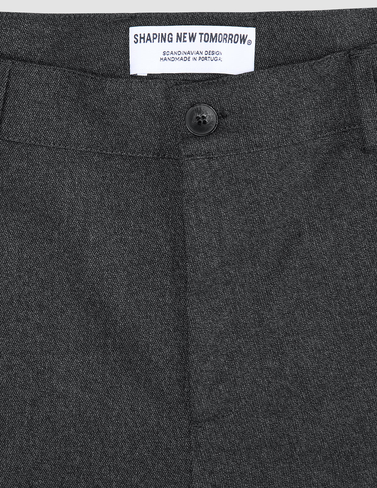 Close-up of the waistband and button of Atelier Pants Relaxed Dark Grey, featuring a white interior label that reads SHAPING NEW TOMORROW, SCANDINAVIAN DESIGN, HANDMADE IN PORTUGAL.