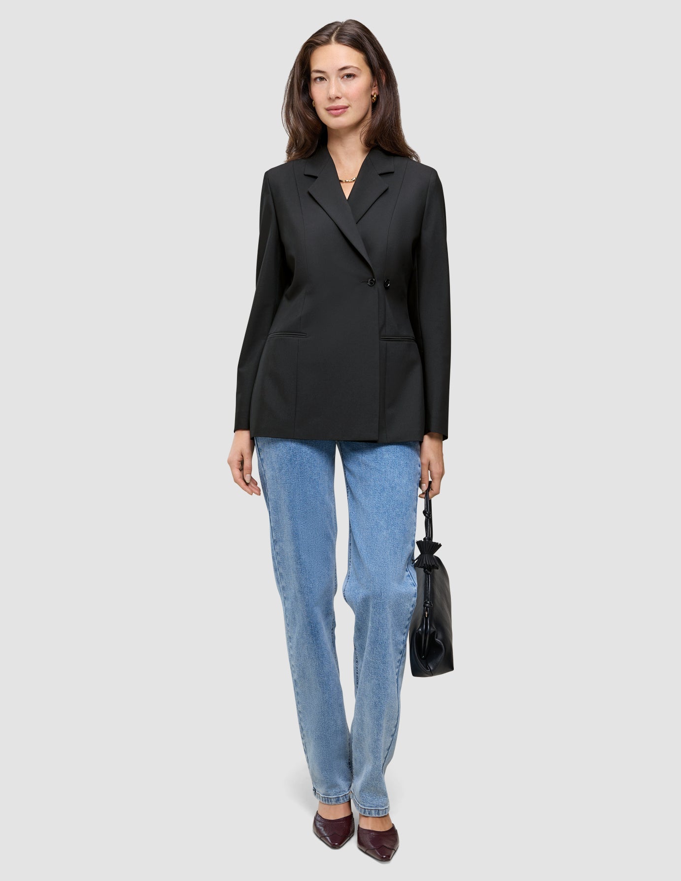 A woman wearing a black Women’s Avira Blazer Black, blue jeans, and dark shoes, holding a small black handbag.