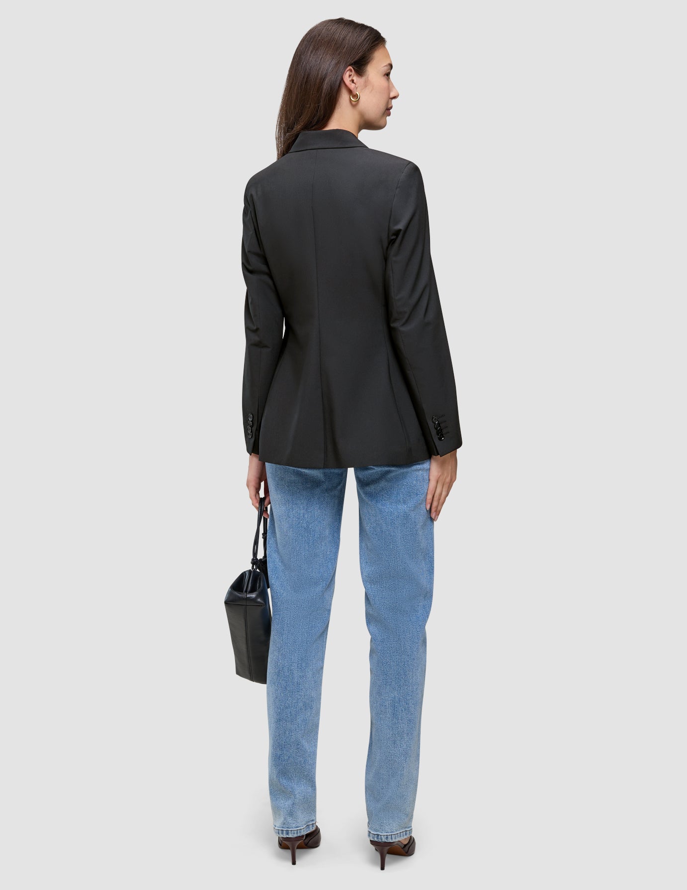 A woman seen from behind wearing a Women’s Avira Blazer Black, light wash jeans, and heeled shoes, carrying a black handbag.