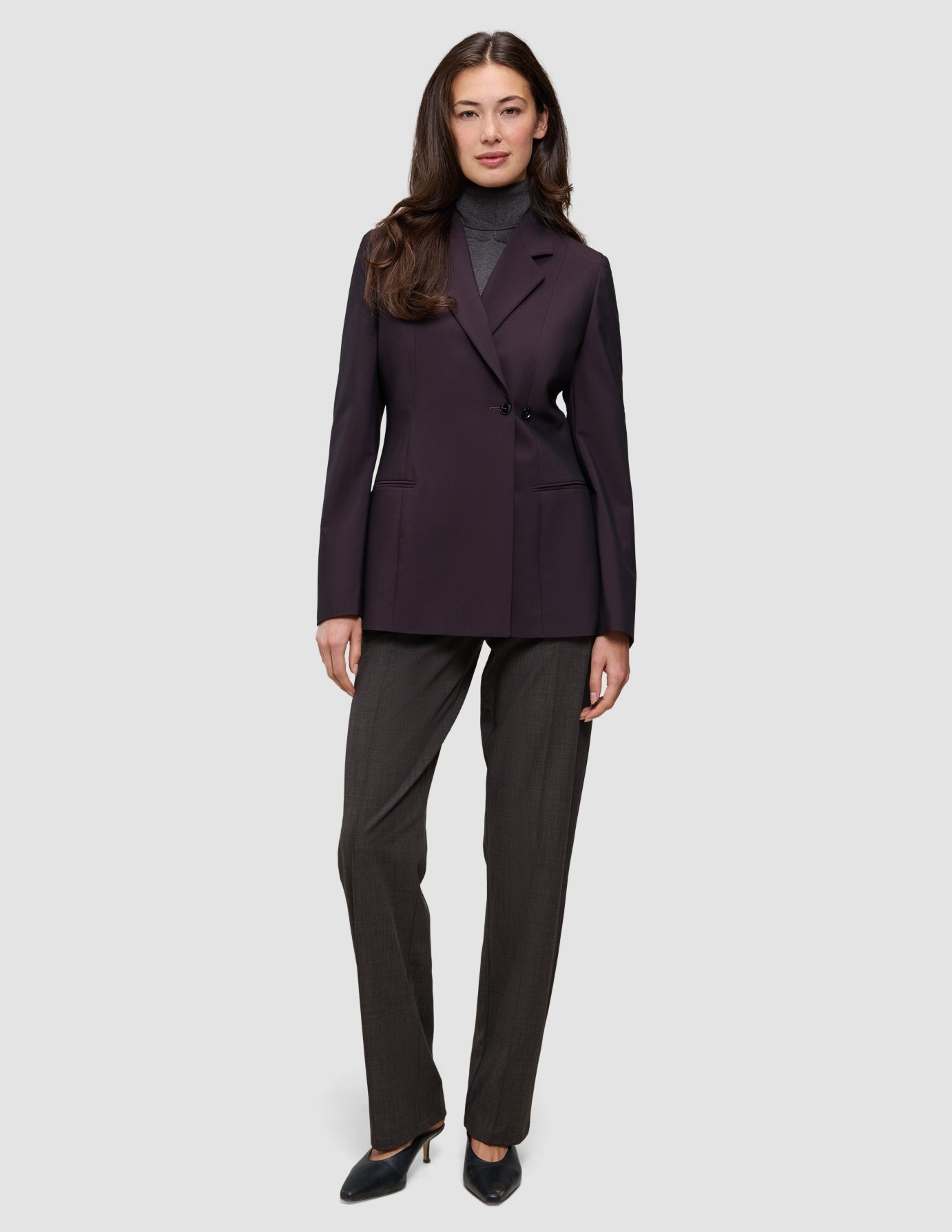 A woman wears a dark burgundy Women’s Avira Blazer Burgundy Berry over a gray turtleneck and dark trousers with black heels, embodying a sleek Scandinavian design.