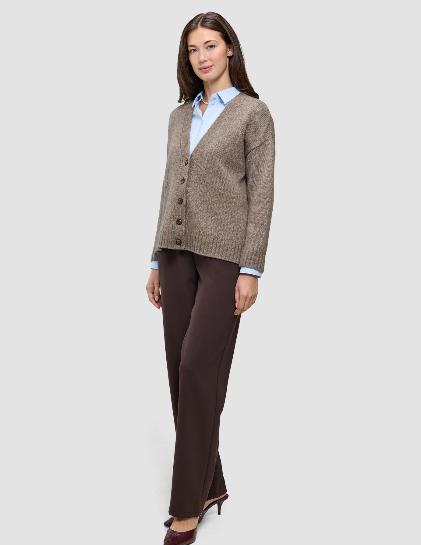 A woman wearing a Women’s Chunky Soft Wool Knit Cardigan Taupe Melange over a light blue collared shirt and dark brown trousers.