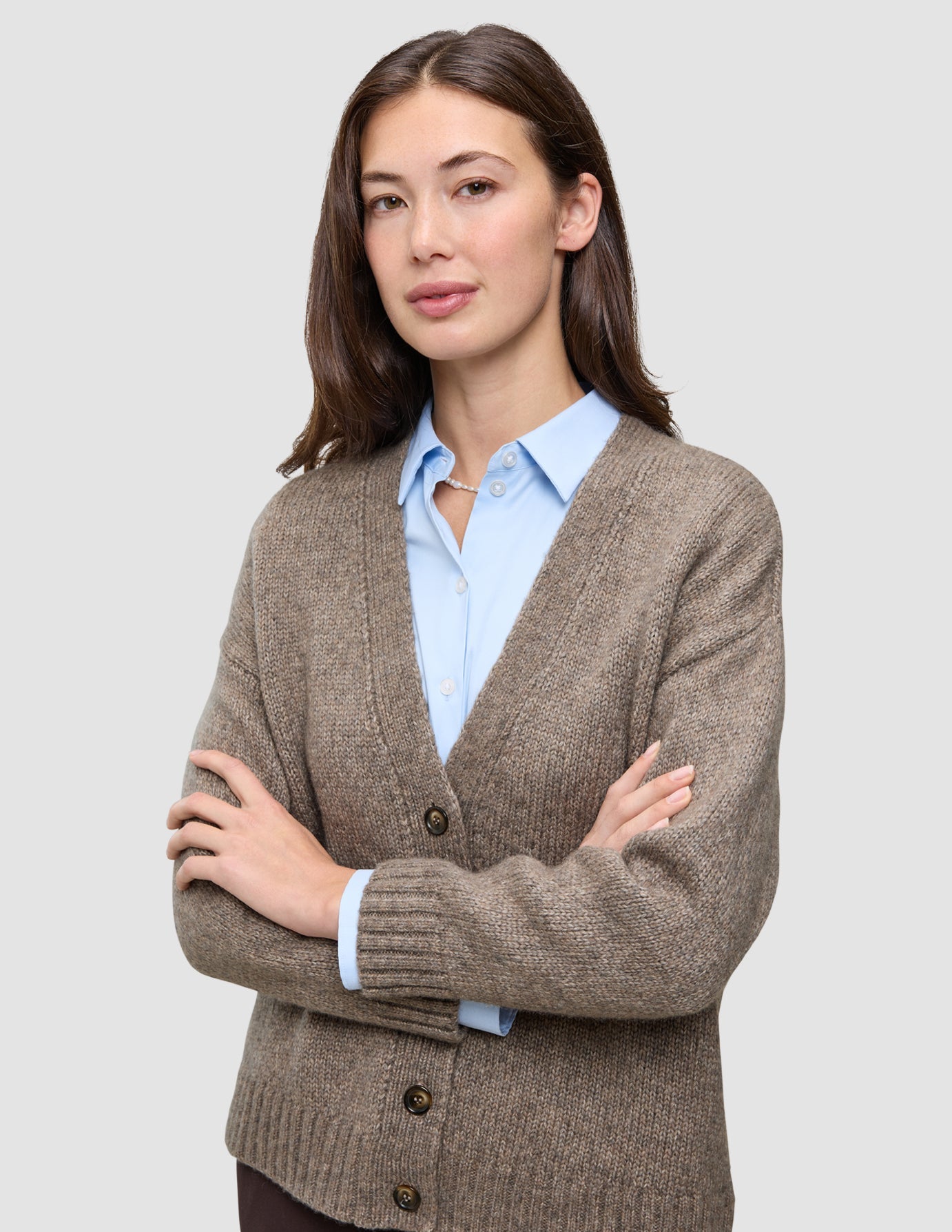 A woman with her arms crossed wears a cozy, taupe, V-neck Women’s Chunky Soft Wool Knit Cardigan Taupe Melange over a light blue button-down shirt.