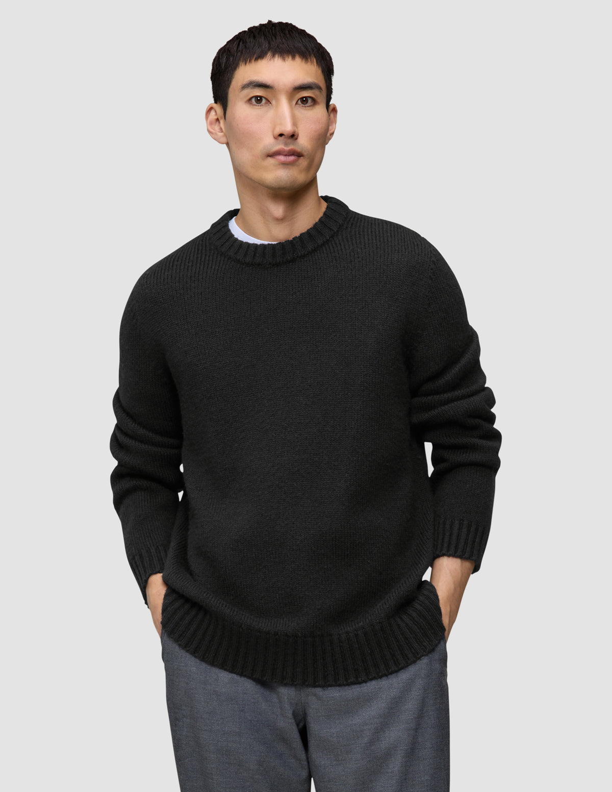 A man wears a black Chunky Soft Wool Knit Crewneck sweater with a thick ribbed collar and cuffs, with his hands in his pockets.
