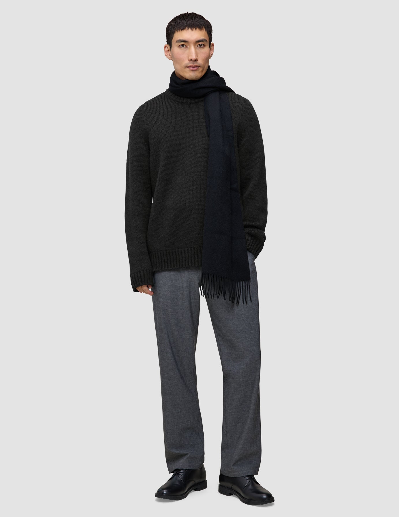 A man wearing a Chunky Soft Wool Knit Crewneck Black, a dark scarf, grey trousers, and black shoes.